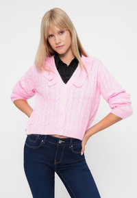 Ροζ πλεκτό κασκόλ με V-λαιμό, σχέδιο καλωδίου και κοντά μανίκια, διαθέτει πέντε κουμπιά και λεπτομέρεια για γιακά. Συνδυάζεται με σκούρα τζιν.