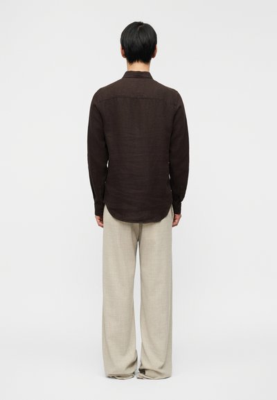 Καφέ λινό πουκάμισο με γιακά που κουμπώνει, μακριά μανίκια και στρογγυλεμένη κάτω ραφή, συνδυασμένο με ανοιχτό μπεζ παντελόνια με φαρδιά μπατζάκια.