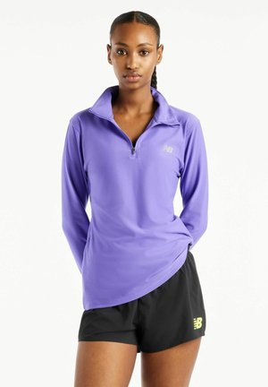 Slank kvinde med fletninger har en lilla quarter-zip trøje og sorte sportsshorts på, og står med hænderne bag ryggen.