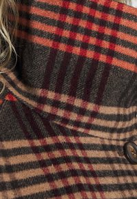 Primer plano de una tela de cuadros en tonos marrón, rojo y negro, con un botón oscuro y mechones de cabello rubio visibles en el lado izquierdo.