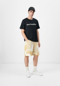 Juoda marškinėliai su baltu "marimekko" tekstu, geltonos šortai su dideliais raštais, balti sportbačiai ir juoda kepurė.