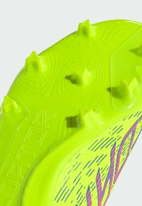 Suela de bota de fútbol verde brillante, texturizada con clavos en relieve. Presenta la marca "ADIDOR" y líneas de acento vibrantes en rosa y azul.