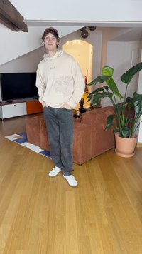 Hoodie bege claro com detalhe gráfico, jeans cinza escuro e tênis brancos. Uma sala de estar com um sofá de veludo cotelê marrom e uma planta verde.
