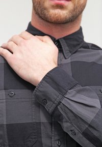 Schwarz-graues kariertes Hemd mit Button-Down-Kragen, mit strukturiertem Stoff und sichtbaren Nähten an Manschetten und Knopfleiste.