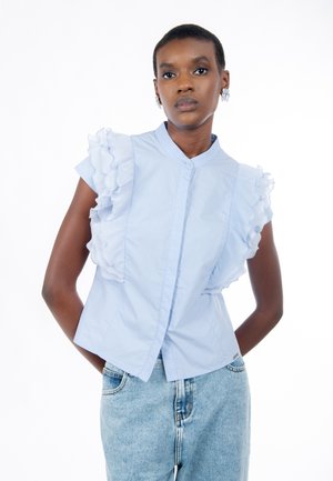 Hellblause Kurzarmbluse mit Rüschenakzenten an den Schultern, vorne mit Knöpfen versehen und einem strukturierten Design. Lässiger Look mit Jeans.