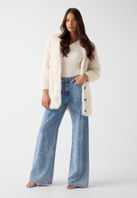 Fuzzy witte cardigan over een licht shirt, gecombineerd met high-waisted, wijde, lichtblauwe spijkerbroek. Naden en knoopdetails zichtbaar.