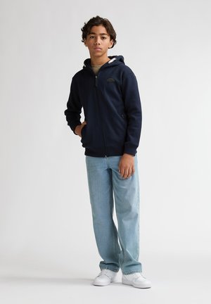 Hanorac bleumarin cu fermoar, glugă cu căptușeală albă, buzunare frontale și un logo pe piept, asortat cu blugi deschis la culoare și pantofi albi.