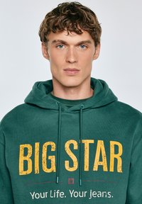 Zelen pulover s kapuco in vrvico, ki ima velik rumen napis "BIG STAR" ter manjši napis spodaj. Mehka tkanina, sproščen kroj, brez vidnih vzorcev.