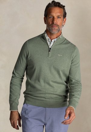 Groene sweater met ritssluiting in zachte stof. Heeft ribgebreide manchetten en onderrand, gelaagd over een patronen shirt. Model draagt lichtblauwe broeken.