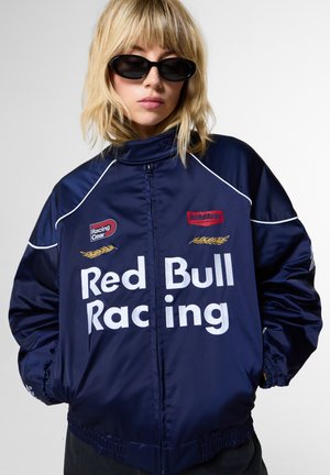 Persona con cabello rubio lleva gafas de sol ovaladas negras y una chaqueta azul marino de Red Bull Racing con texto blanco y parches.