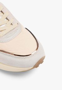 Primer plano de la punta de una zapatilla beige con ribete de ante, detalle metálico brillante, cordones a juego y suela de goma marrón claro sobre fondo blanco.