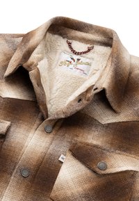 Giacca a quadri marrone e beige con fodera in pile crema, bottoni a pressione metallici, tasche sul petto e etichetta del marchio all'interno del colletto.