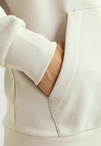 Sudadera color beige claro con una textura suave, puños acanalados y un bolsillo frontal. Se pueden ver costuras a lo largo de los bordes para añadir estructura.