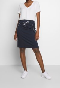 Ein navyblauer Jerseykilt mit einem Kordelzugbund und zwei seitlichen Reißverschlusstaschen, kombiniert mit einem kurzärmeligen weißen V-Ausschnitt-Shirt und weißen Turnschuhen.