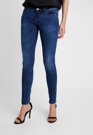 Femme portant un jean skinny bleu foncé et des sandales noires à talons hauts, debout devant un fond clair uni.