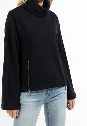 Vrouw die een zwarte coltrui met lange mouwen en zijritsen draagt en een lichtblauwe spijkerbroek, staand tegen een witte achtergrond.