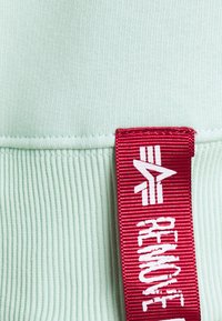 Tessuto verde menta chiaro con una texture a righe; presenta un'etichetta rossa prominente con la scritta "RIMUOVERE" stampata in bianco, attaccata alla cucitura.