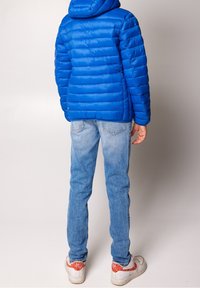 Chaqueta acolchada azul con paneles cosidos, capucha y puños elásticos, combinada con jeans azul claro y zapatillas blancas con acentos rojos.