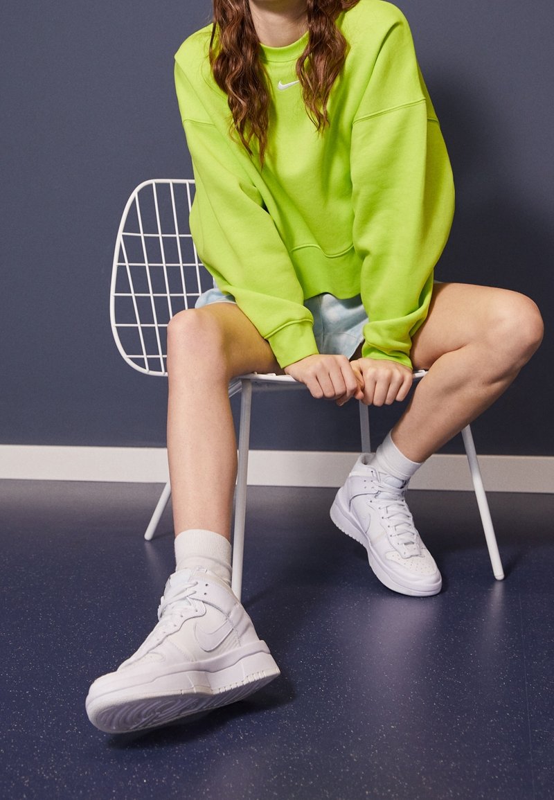 Bílé kotníkové tenisky z texturované kůže, kulatá špička a polstrovaný límec. N nošené s bílými ponožkami a kombinované s jasně zelenou crop mikinou.