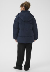 Moteris su garbanotais plaukais, apsirengusi tamsiai mėlynu pūkiniu švarku su kapišonu, juodomis plačiomis kelnėmis ir juodais batais, atsukta nugara prieš vientisą foną.