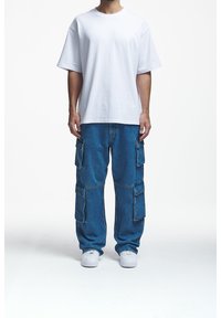 Muž v jednoduchém bílém oversized tričku, volných modrých denimových cargo kalhotách s několika kapsami a bílých teniskách, stojící na bílém pozadí.