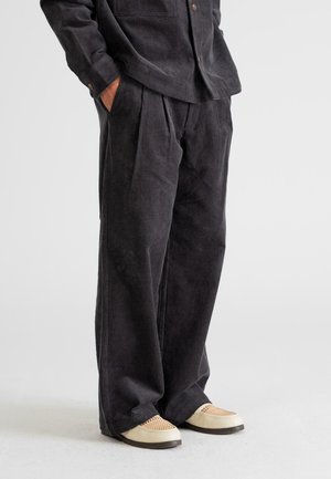 Black corduroy pants with a relaxed fit, featuring front pleats and a soft texture. Paired with light-colored flat shoes.