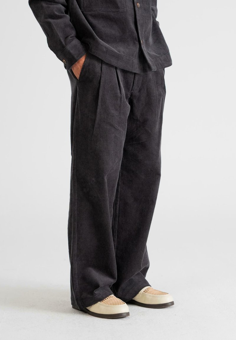 Black corduroy pants with a relaxed fit, featuring front pleats and a soft texture. Paired with light-colored flat shoes.