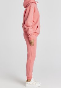 Persona de pie de lado, vistiendo una sudadera rosa, joggers a juego y zapatillas blancas, contra un fondo blanco liso.