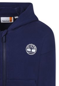 Sweat à capuche zippé bleu marine avec un logo d'arbre blanc sur la poitrine, doté d'une capuche et de poches avant. Tissu doux avec une texture lisse.