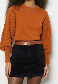Pull orange rouille avec manches texturées et à motifs, associé à une jupe en velours côtelé noire et une ceinture marron cloutée. Accessoire : colliers argentés superposés.