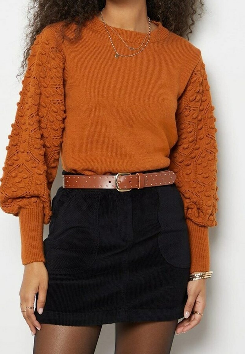 Pull orange rouille avec manches texturées et à motifs, associé à une jupe en velours côtelé noire et une ceinture marron cloutée. Accessoire : colliers argentés superposés.