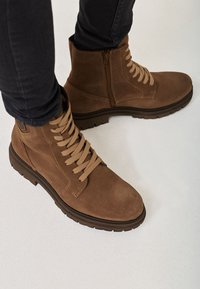 Stivaletti alla caviglia in suede marrone con lacci beige e cerniera laterale. Suola robusta, tacco piatto e dettagli di cucitura sulla tomaia.