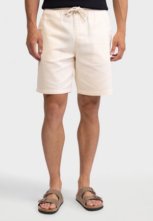 Mies, joka pukeutunut kermaisiin narushortseihin ja ruskeisiin kaksihihnaisiin sandaaleihin, seisoo yksinkertaista vaaleaa taustaa vasten.