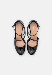 Chaussures en cuir verni noir avec un design à bout fermé, ornées de brides croisées autour de la cheville et d'une finition lisse et brillante.