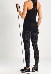 Nainen mustissa harjoitusvaatteissa pitää vastuskumpparia, seisoo valkoista taustaa vasten selkä kameraan päin.