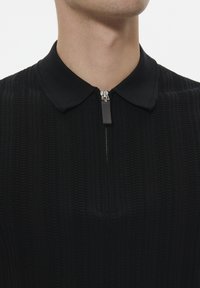 Black knitted polo top featuring a zip neckline, textured fabric, and a structured collar. Close-up view of the upper garment.