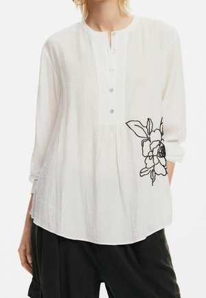 Weiße Bluse mit langen Ärmeln aus strukturiertem Stoff, mit einer Knopfleiste mit drei Knöpfen und schwarzer Blumenstickerei auf der linken Seite, getragen zu schwarzen Hosen.