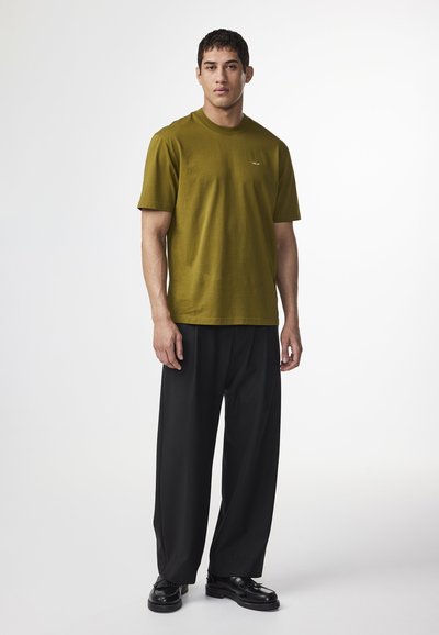 Oliven grøn bomulds t-shirt med korte ærmer, afslappet pasform; båret med løse sorte bukser og sorte sko, minimal branding på brystet.