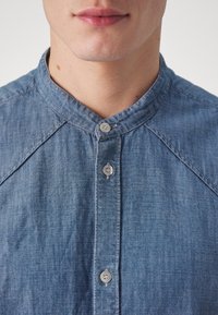 Blå denimskjorta med bandkrage och silverknappar, med en strukturerad tyg och en rak skärning.