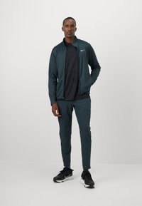 Aktiivne dressipluus tumedas turkoosis, sisaldab jäika jakki ja sobivaid pükse, koos mustade spordikingadega. Nike logo on riietusel nähtav.