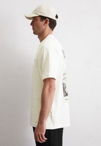 Light beige cotton t-shirt featuring short sleeves and a graphic print on the back. A coordinating beige baseball cap completes the outfit.