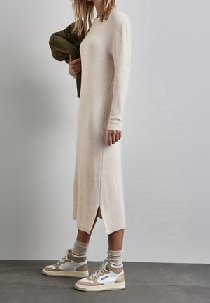 Robe en tricot beige à côtes longues avec fentes sur les côtés, assortie à des baskets montantes blanches et beiges et des chaussettes légères grises à motifs.