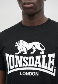 Persoană purtând un tricou negru cu un logo alb cu un leu care răcnește și textul „LONSDALE LONDON” pe piept.