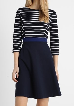 Vrouw draagt een marineblauwe A-lijn rok en een zwart-wit gestreepte longsleeve met een blauwe tailleband.