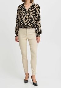 Blouse à motif noir et beige avec ourlet froncé élastiqué, manches longues et col en V, associée à un pantalon beige ajusté et des talons noirs.