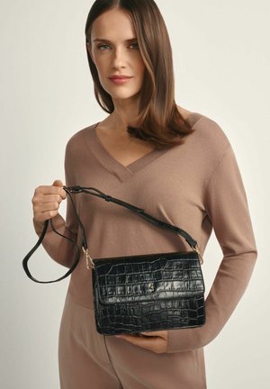 Kobieta w beżowym swetrze z dekoltem w serek, trzymająca czarną torebkę na ramię o fakturze krokodyla, z klapką i regulowanym paskiem.