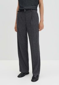 Pantalons gris à jambes larges avec une finition texturée, dotés de plis avant et d'une ceinture noire. Accompagnés de chaussures noires.