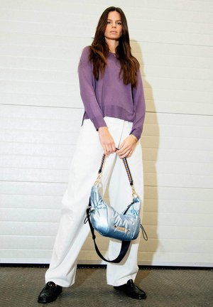 Femme aux longs cheveux bruns tenant un sac à main bleu métallique, portant un pull violet, un pantalon blanc à jambes larges et des chaussures noires devant un mur blanc.