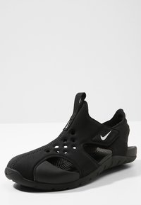 Sandalia deportiva negra de Nike con diseño tipo slip-on, parte superior transpirable con perforaciones, suela texturizada y un logo blanco en contraste.