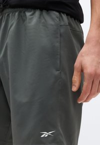 Κοντινό πλάνο ενός ατόμου που φορά σκούρα γκρίζα αθλητικά σορτς με ελαστική μέση και λευκό λογότυπο Reebok κοντά στο τελείωμα.